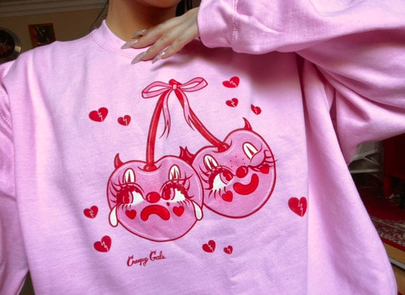 Pink cotton and polyester crew neck sweat shirt featuring pink and red clown cherries with devil horns with one face sad and crying and other happy and winking. The cherries character is surrounded by red hearts