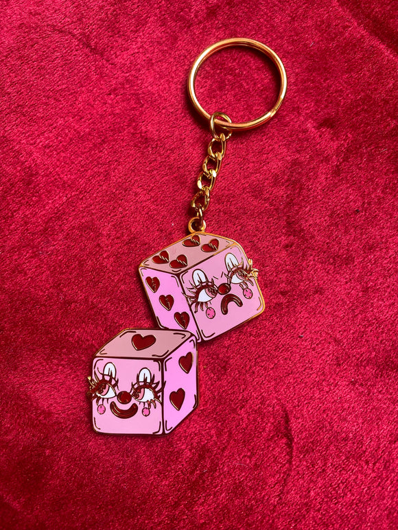 Gold keychain of a pink pair of dice with red hearts . The face i the front is happy the one in the back is angry  