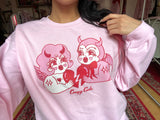 Pink cotton and polyester unisex crew neck sweat shirt featuring kitschy pink and white devil cherub and a red devil holding each side of a red broken heart.