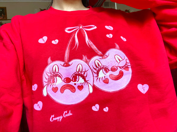 Red cotton and polyester unisex crew neck sweat shirt featuring kitschy pink devil cherries with clown makeup on. One cherry crying and the other winking. They are tied together with a pink bow and surrounded by pink broken hearts