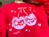 Red cotton and polyester unisex crew neck sweat shirt featuring kitschy pink devil cherries with clown makeup on. One cherry crying and the other winking. They are tied together with a pink bow and surrounded by pink broken hearts