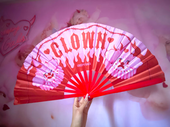 Large pink and red hand fan with red bamboo handle and silk fan. It features the word clown in the center with Poopywise the clown on each size and red flames in the background and red Barb wire on top