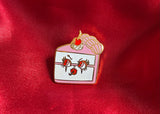 Gold plated enamel pin of a kitschy slice of cake with a cherry on top and has a face smoking