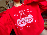 Red cotton and polyester unisex crew neck sweat shirt featuring kitschy pink devil cherries with clown makeup on. One cherry crying and the other winking. They are tied together with a pink bow and surrounded by pink broken hearts